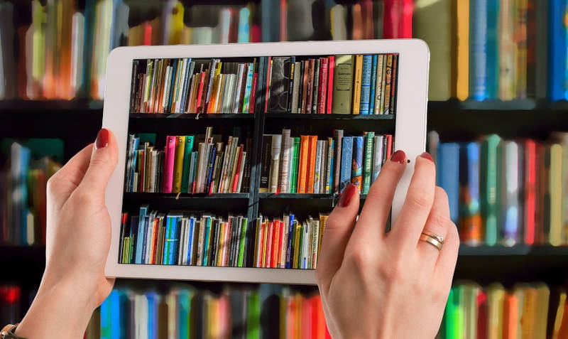 Ein Tablet vor einem Bücherregal. Der Bildschirm des Tablets zeigt die dahinter stehenden Bücher.