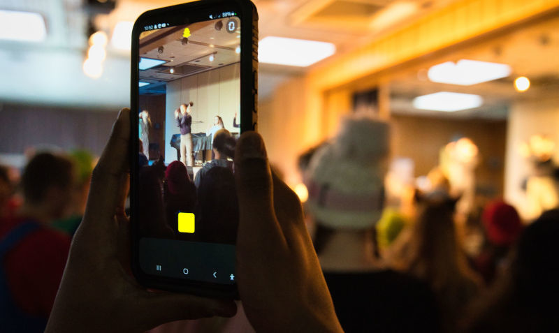 Ein hochgehaltenes Handy im Fotomodus zeigt eine Karnevalsfeier.